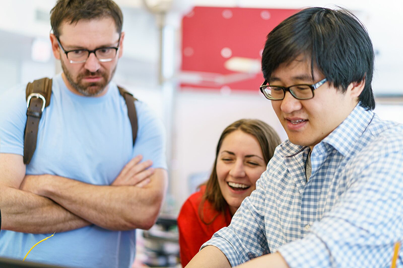 Three people gather around a common project. One is talking, one smiling, and the third is watching the (unseen) project closely.