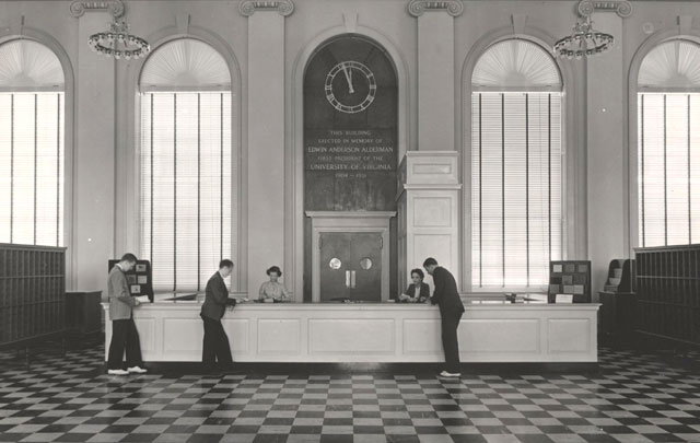 A vintage photo from the main library's early days when there was a central front desk. Three men are at the desk talking with librarians.