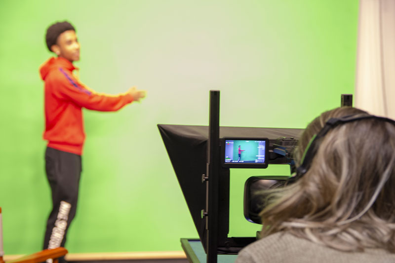 Man against a "green-screen" with a woman working at the camera.